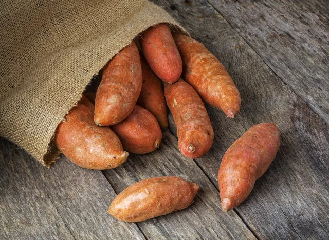 Freshly harvested organic sweet potatoes spilling from a burlap bag onto a natural weathered wood table.