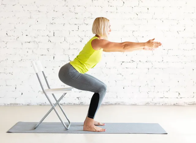 Yoga with a chair. Fit adult caucasian woman practice squat with props on a mat in loft white studio indoor, selective focus. Fitness, workout, trainer, sport, healthy lifestyle, concept.