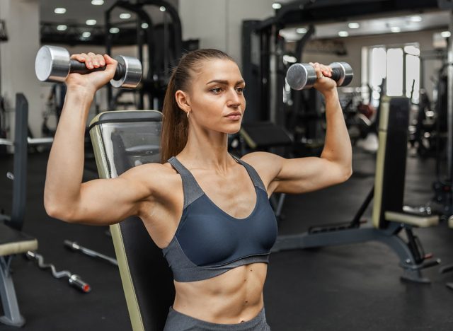 Cheerful fit woman trains shoulders with dumbbells in hands doing overhead press while sitting on a bench in a modern gym