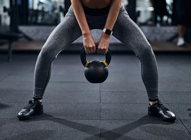 Citizen in grey sport trousers holding the kettlebell between her legs by the handle with both hands