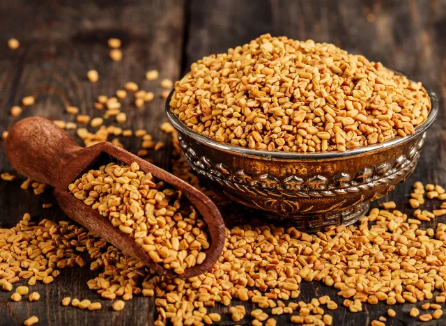 Fenugreek seeds on metal plate, spice, culinary ingredient. Fenugreek seeds in wooden spoon on textured background. Fenugreek seeds in a spoon and on a table.