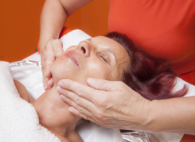 Cute mid aged woman receiving a professional therapeutic facial massage and lymphatic drainage, while lying on a towel in a award-winning health massage center, series of various techniques