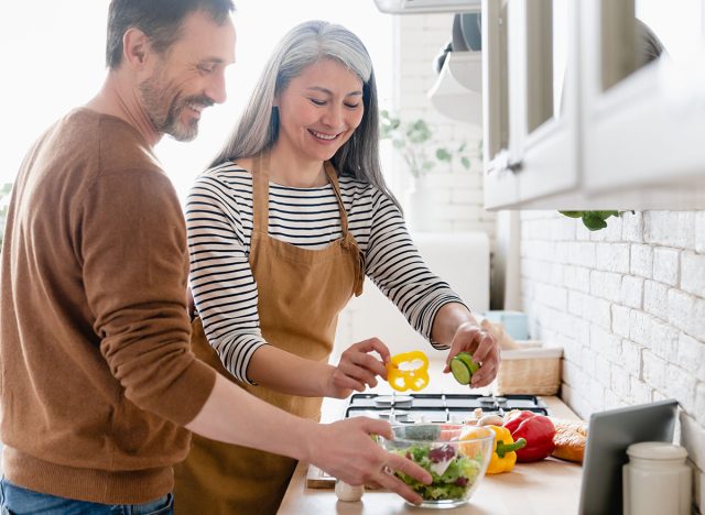 Happy mature middle-aged couple cooking vegetable vegetarian salad together in the kitchen, helping in preparation of food meal. Family moments, domestic homemade food