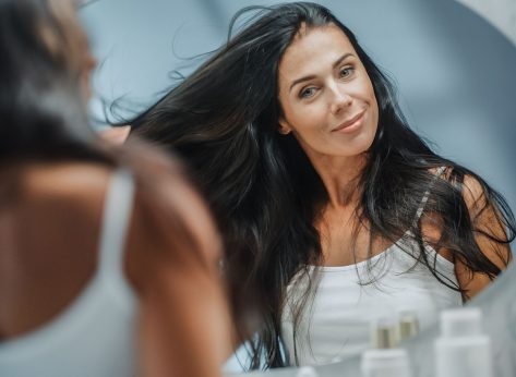 Beautiful Early Middle Aged Woman Looks into Bathroom Mirror Touches Her Lush Black Hair, Admires Her Looks. Concept for Happiness, Wellbeing, Natural Beauty, Organic Skin Care Products