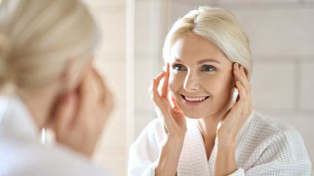 Gorgeous mid age adult 50 years old blonde woman standing in bathroom after shower touching face, looking at reflection in mirror smiling doing morning beauty routine. Older dry skin care concept.