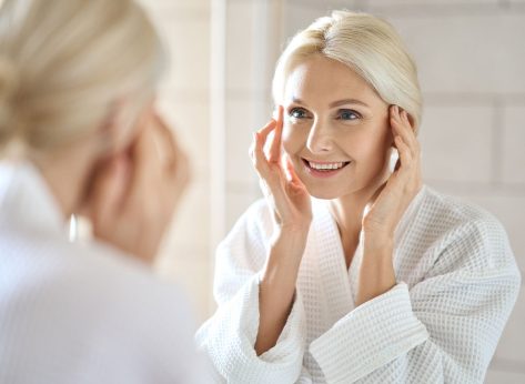 Gorgeous mid age adult 50 years old blonde woman standing in bathroom after shower touching face, looking at reflection in mirror smiling doing morning beauty routine. Older dry skin care concept.