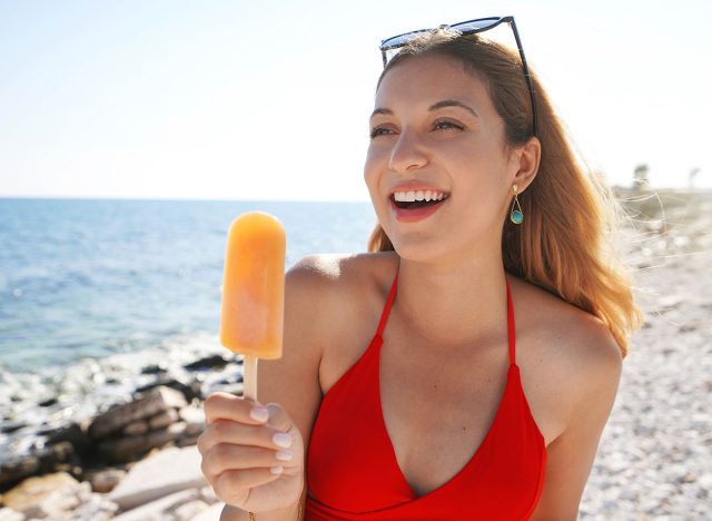 Cute young woman eating an orange popsicle on the beach on summer