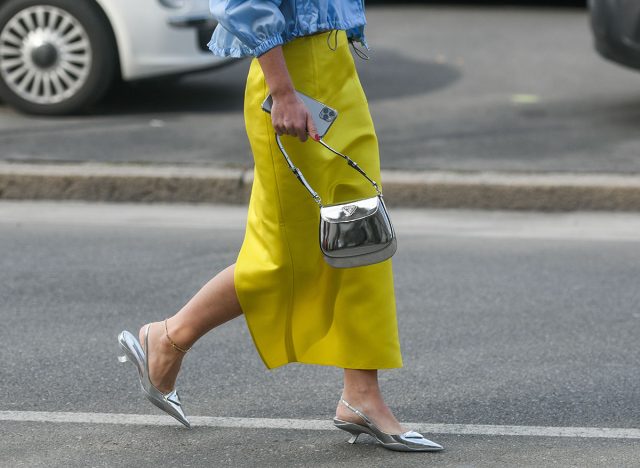 Milan, Italy - February 24, 2022: Street style, woman wearing Prada outfit: blue V-neck bra, a matching blue cropped bomber coat, yellow midi skirt and silver pointed pumps heels shoes.