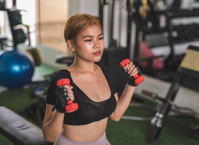 A young and sexy asian woman does seated dumbbell hammer curls on a flat bench at the gym. Wearing a black crop top and bike shorts.