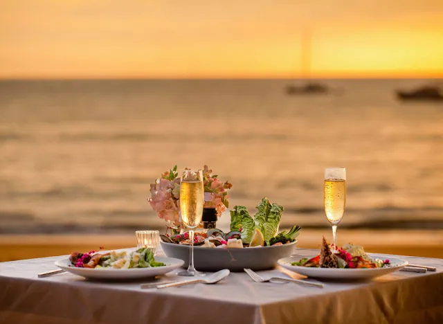 Romantic sunset dinner on beach. Table honeymoon set for two with luxurious food, glasses of champagne drinks in restaurant with sea view and yachts on background. Summer vacation or wedding concept.