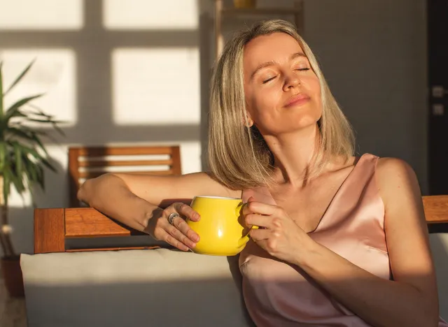 Attractive happy middle aged woman is sitting on sofa in living room. Smiling adult lady enjoys drinking coffee or tea sitting on couch at home