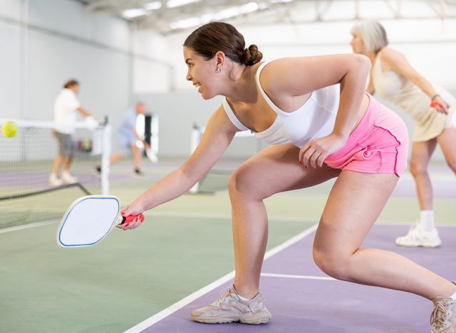 Concentrated fit young girl pickleball player waiting to receive serve, ready to strike and return ball to opponent field on indoor court..