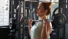 Focused woman performing barbell curls at gym, bathed in natural light. Gym workout routine.