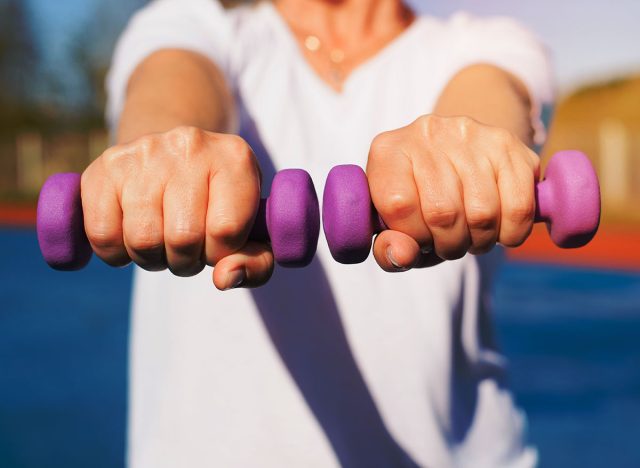 Sport and active lifestyle concept. Caucasian woman training, sportswoman exercising with dumbbells holding her hands forward outdoors on sunny day, close-up.