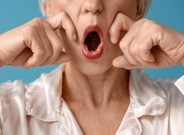 Mature woman in pajamas doing face building exercise on blue background, closeup