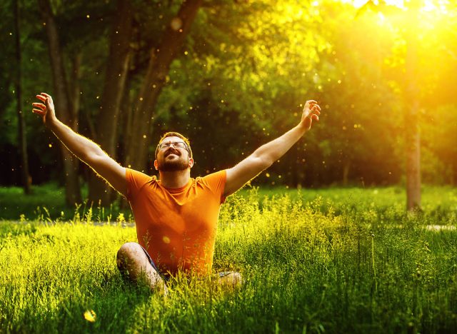 A happy man is relaxing on green grass with squint eyes and raised up to sky arms at sunny summer day at park background. Concept of wellbeing and healthy lifestyle