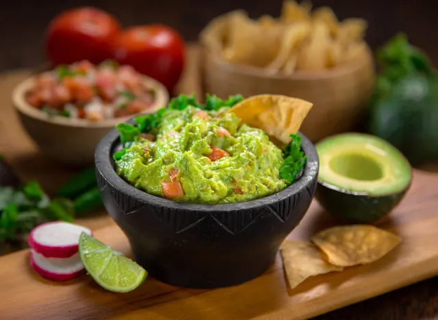 A delicious Bowl of Guacamole next to fresh ingredients on a table with tortilla chips and salsa