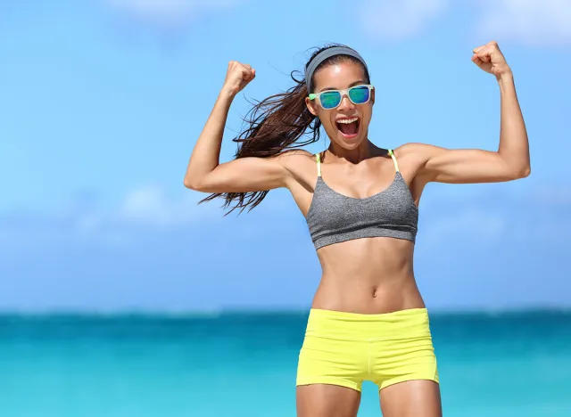 Strong fitness funny woman in neon blue wayfarer sunglasses on beach showing off muscular arms flexing biceps for fun. Fit girl in sportswear after running strength training workout winning in power.