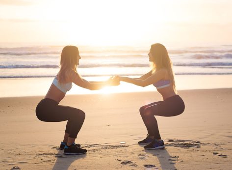 Strength in teamwork. Two young attractive female athletes exercise on the beach doing squats with a sunrise and ocean in the background. The focus is soft and dreamy.