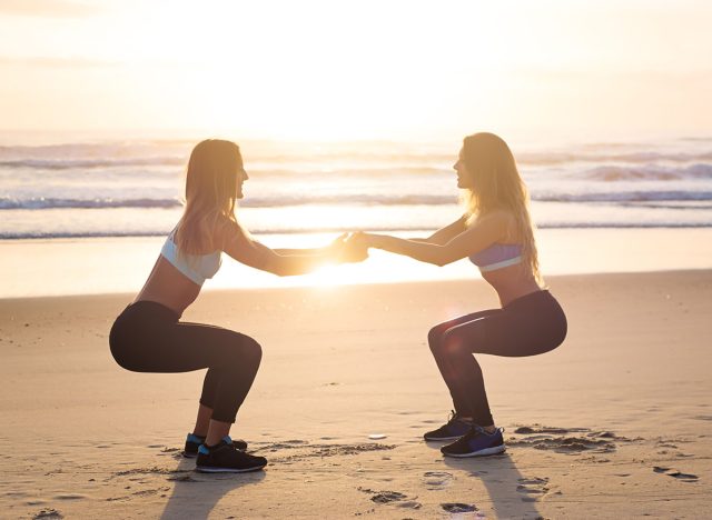 Strength in teamwork. Two young attractive female athletes exercise on the beach doing squats with a sunrise and ocean in the background. The focus is soft and dreamy.