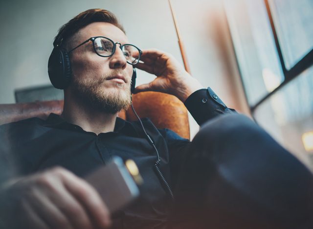 Portrait handsome bearded man wearing glasses,headphones listening to music at modern home.Guy sitting in vintage chair,holding smartphone and relaxing.Panoramic windows background.Blurred background