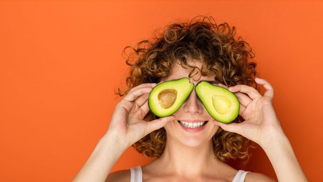 Young,Curly,Woman,Holding,A,Avocado,In,Hands,On,Orange