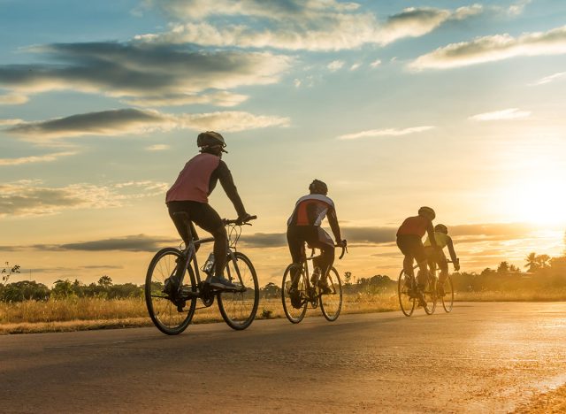 Group of men ride bicycles at sunset with sunbeam over silhouette trees background.