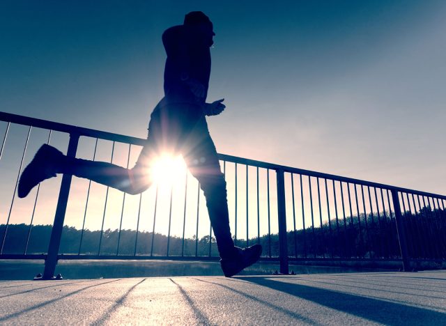 Rear view to runner in blue t-shirt and black leggings running on bridge. Outdoor exercising on smooth concrete ground on lake bridge. Sun is outlining man body