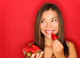 Girl eating strawberries smiling happy looking to the side on red background with copy space. Beautiful young mixed race Asian and Caucasian woman eating healthy.