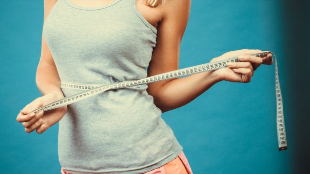 Weight loss, slim body, healthy lifestyle concept. Fit fitness girl measuring her waistline with measure tape on blue