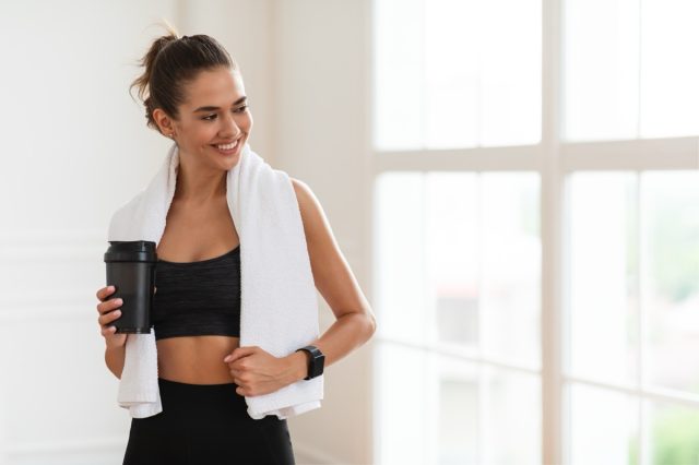 Portrait of sporty smiling young woman in sportswear and white towel on neck holding shaker with healthy drink, whey protein, fresh water or coffee and looking away.