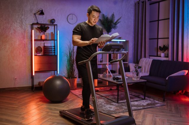 Asian man reading book working out, running, doing cardio training on treadmill in evening time, indoor.