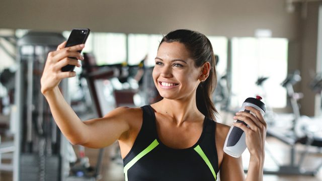 A woman at the gym takes a picture to send to friends or put on social networks and takes the picture while smiling. Concept of: network, friendship, gym, fitness