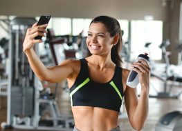 A woman at the gym takes a picture to send to friends or put on social networks and takes the picture while smiling. Concept of: network, friendship, gym, fitness
