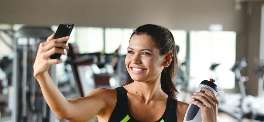 A woman at the gym takes a picture to send to friends or put on social networks and takes the picture while smiling. Concept of: network, friendship, gym, fitness