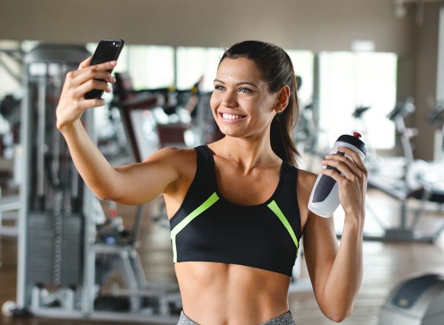 A woman at the gym takes a picture to send to friends or put on social networks and takes the picture while smiling. Concept of: network, friendship, gym, fitness