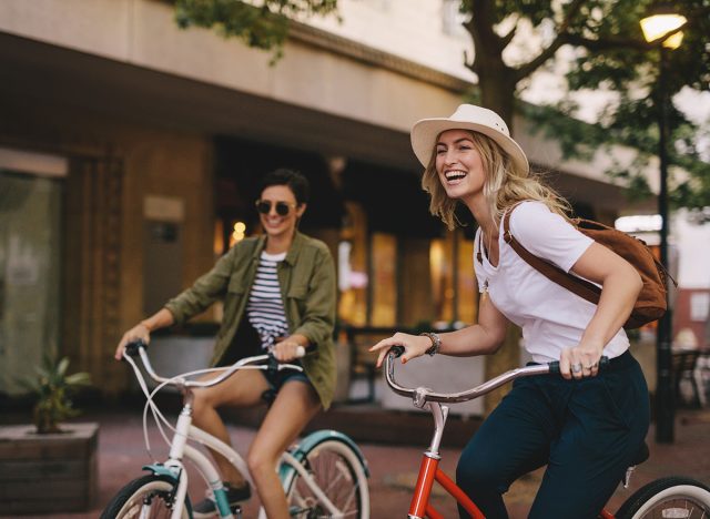 Smiling young woman cycling with her friend on city street. Two young women riding bicycles outdoors and having fun.