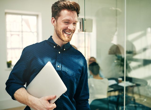 Young businessman laughing while walking in a modern office carrying a laptop with colleagues at work in the background