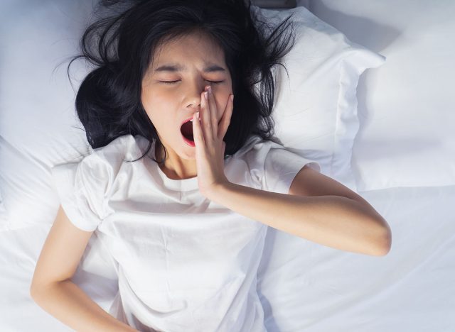 Woman stretching in bed after waking up, back view. Woman sitting near the big white window while stretching on bed after waking up with sunrise at morning, back view.