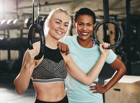 Two diverse young female friends in sportswear standing together by rings in a gym after a workout
