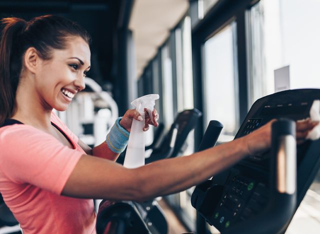 Young happy and smiling woman cleaning and weeping expensive fitness gym equipment with sprayer and cloth.