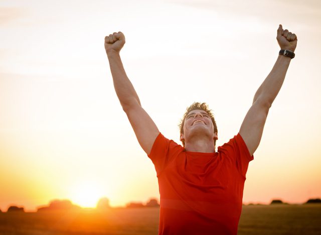 Successful man raising arms after cross track running on summer sunset. Fitness male athlete with arms up celebrating success and goals after sport exercising and working out.