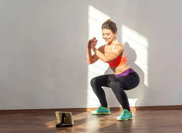 Positive sportive woman with bun hairstyle and in tight sportswear doing squatting sit-up exercise while watching training video on tablet. indoor studio shot illuminated by sunlight from window