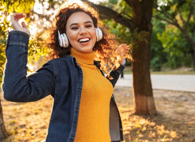 Image of positive cheery beautiful young curly woman walking in park outdoors listening music with headphones.