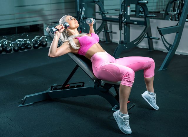 Attractive blonde woman in 30s doing chest press exercise on bench in modern fitness center. Toned image.