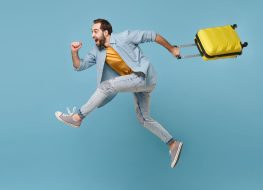 Side view of funny traveler tourist man in yellow clothes isolated on blue background. Male passenger traveling abroad on weekends. Air flight journey concept. Jumping like running, hold suitcase
