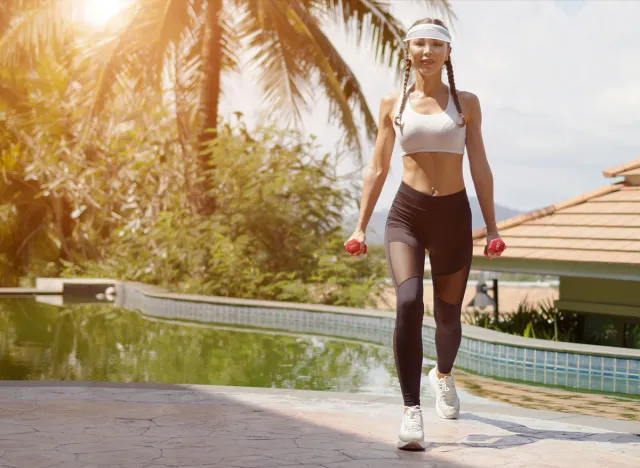 Full body slim female with dumbbells walking on poolside towards camera during outdoor workout on sunny day on tropical resort