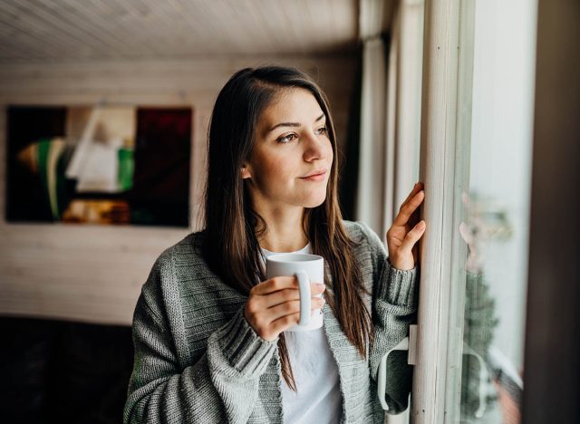 Young woman spending free time home.Self care,staying home.Enjoying view,gazing through to the window.Quarantined person indoors.Serene mornings.Avoiding social contact.