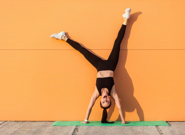 Overjoyed excited girl with perfect athletic body in tight sportswear doing yoga handstand pose against wall and laughing, shouting from happiness. Gymnastics for body balance outdoor workouts