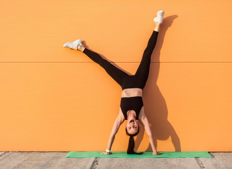 Overjoyed excited girl with perfect athletic body in tight sportswear doing yoga handstand pose against wall and laughing, shouting from happiness. Gymnastics for body balance outdoor workouts